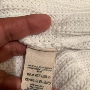 May include: Close-up of a white knit fabric with a tag attached. The tag has text in multiple languages, including English, and care symbols. The fabric appears to be made of cotton.