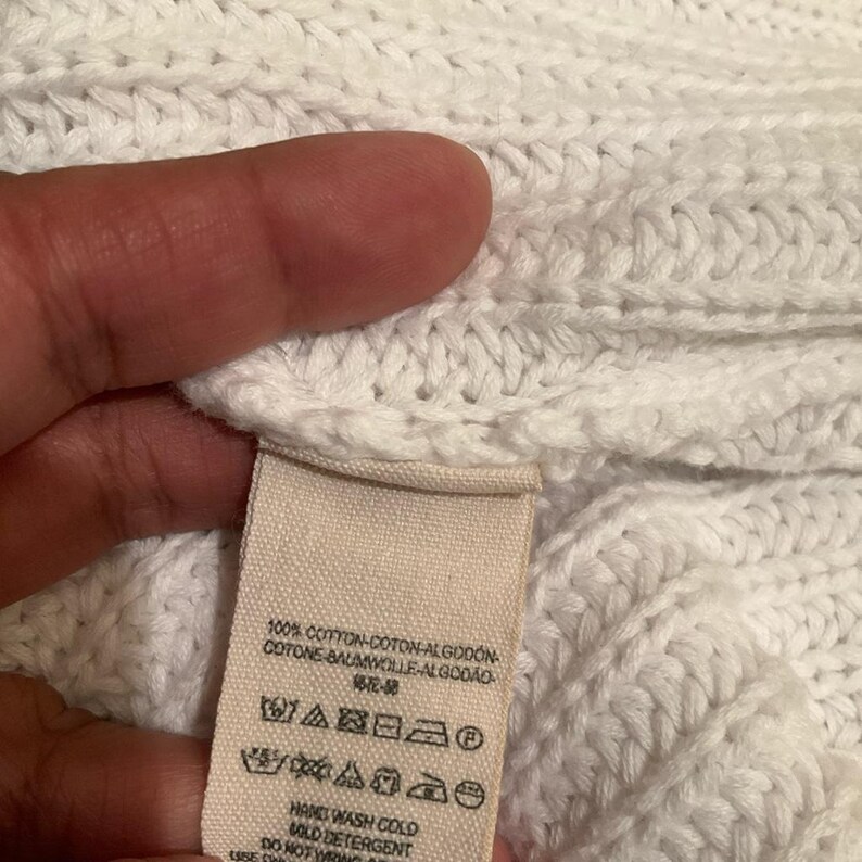 May include: Close-up of a white knit fabric with a tag attached. The tag has text in multiple languages, including English, and care symbols. The fabric appears to be made of cotton.