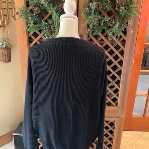 Pode incluir: Um su&eacute;ter de malha canelada azul marinho exibido em um manequim. O su&eacute;ter tem gola redonda e mangas compridas. O fundo inclui um arm&aacute;rio de madeira com design de treli&ccedil;a e grinaldas decorativas.