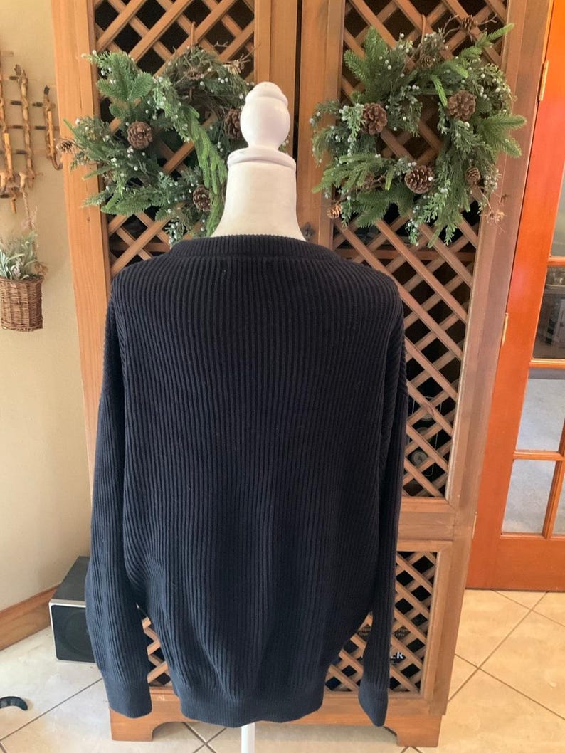 Pode incluir: Um su&eacute;ter de malha canelada azul marinho exibido em um manequim. O su&eacute;ter tem gola redonda e mangas compridas. O fundo inclui um arm&aacute;rio de madeira com design de treli&ccedil;a e grinaldas decorativas.