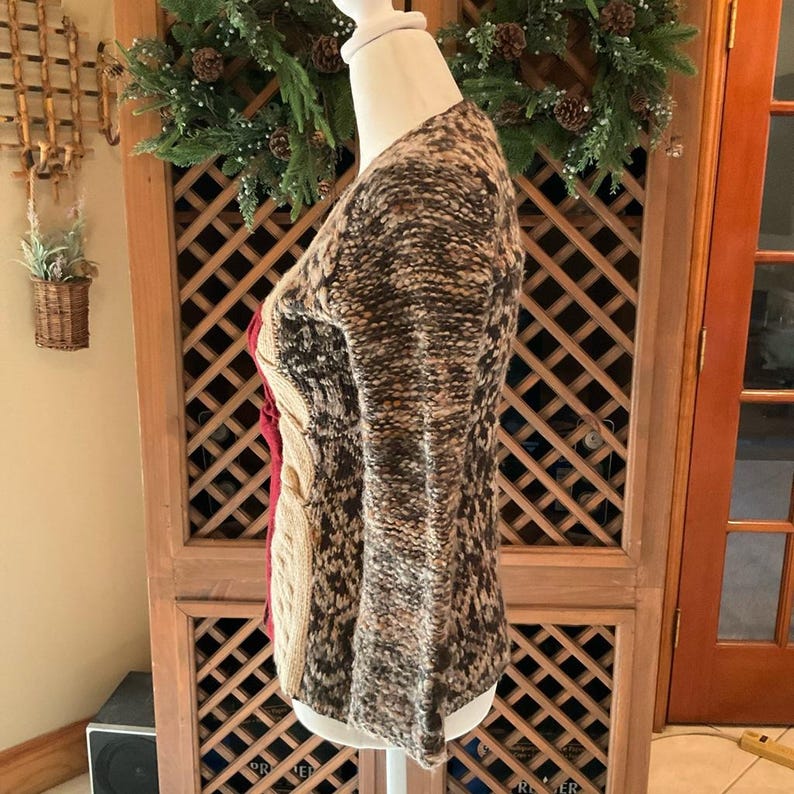 May include: A multi-colored sweater with a cable knit design. The sweater features a mix of brown, beige, and red hues. The sleeves are long and flared. The sweater is displayed on a mannequin.