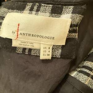 May include: Close-up of a tag from Anthropologie. The tag is cream-colored with black text and a red vertical line. The tag reads "by ANTHROPOLOGIE" and "made in india fabrique en inde". The tag also shows UK 8, US 4, and EU 36.