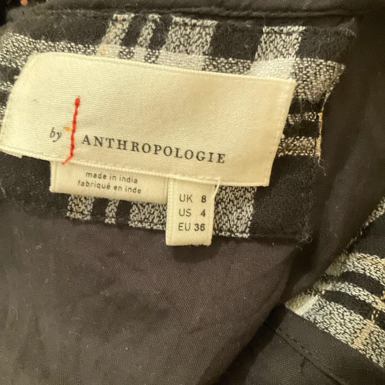 May include: Close-up of a tag from Anthropologie. The tag is cream-colored with black text and a red vertical line. The tag reads "by ANTHROPOLOGIE" and "made in india fabrique en inde". The tag also shows UK 8, US 4, and EU 36.