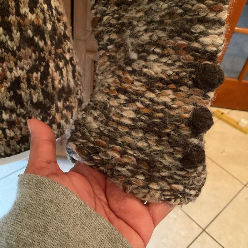 May include: Close-up of a textured, multi-colored knit jacket sleeve. The sleeve features a mix of brown, beige, and gray yarns, with three dark brown, round buttons. The jacket appears to be a warm, textured garment.