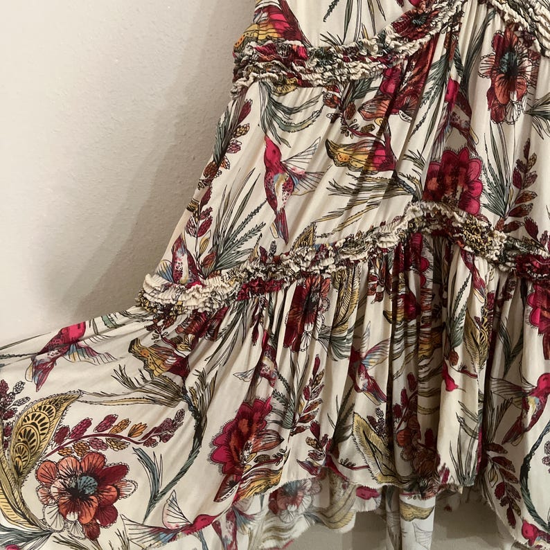 May include: Cream-coloured fabric with a vibrant floral and hummingbird print. The design features red, pink, and yellow flowers, along with green leaves and hummingbirds. The fabric has a ruffled trim.