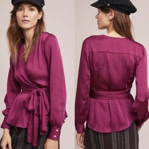 Puede incluir: Top envolvente de manga larga en color magenta intenso. Presenta un escote en V, cierre de lazo en la cintura y pu&ntilde;os abotonados. La tela tiene un brillo sutil, sugiriendo sat&eacute;n o seda. El dise&ntilde;o es elegante y femenino, ideal para ocasiones especiales.