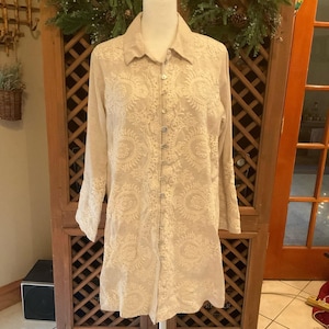 May include: Beige long-sleeve tunic with intricate, embroidered floral and circular patterns. The tunic features a button-down front and a collared neckline. The embroidery is visible on the sleeves and the front of the tunic.