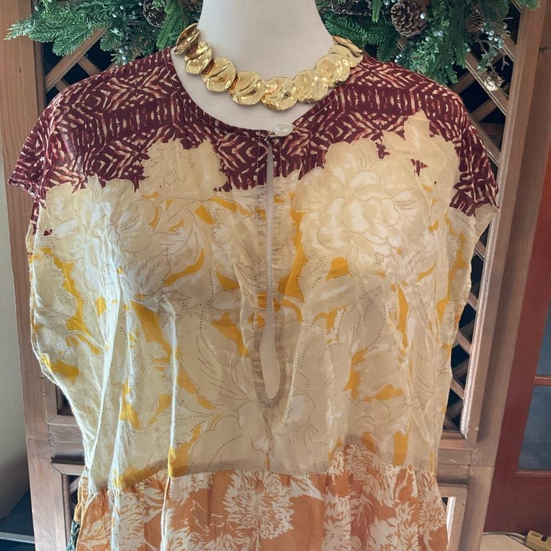 May include: A floral print dress in cream, yellow, and orange, with a burgundy patterned yoke. A gold-toned necklace with circular pendants adorns the neckline. The dress features a loose fit and a keyhole neckline.