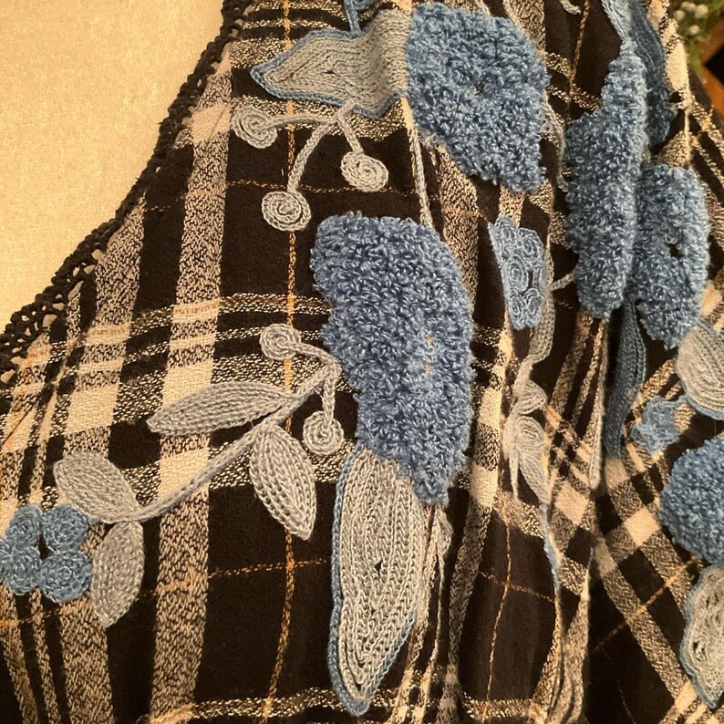 May include: Close-up of a black and white plaid fabric with blue and gray floral embroidery. The embroidery features textured blue flowers and gray leaves, with a crocheted edge.