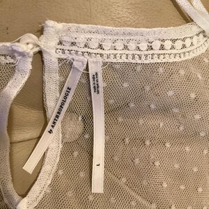 Pu&ograve; includere: Primo piano di un top bianco trasparente con un delicato motivo a pois. Il top presenta uno scollo alto con un bordo in pizzo smerlato. Sono visibili due etichette, una con la scritta "by ANTHROPOLOGIE" e l'altra che indica che il capo &egrave; stato realizzato in India, taglia L.