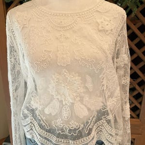 Pu&ograve; includere: Un top in pizzo bianco a maniche lunghe con intricati motivi floreali e geometrici. Il tessuto delicato presenta uno scollo tondo e finiture dettagliate lungo l'orlo e i polsini. Il design trasparente rivela l'indumento sottostante.
