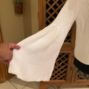 May include: White knit sweater with a flared sleeve. The sleeve is long and bell-shaped, made of a textured knit fabric. The sweater is a solid white color and appears to be a medium weight knit.