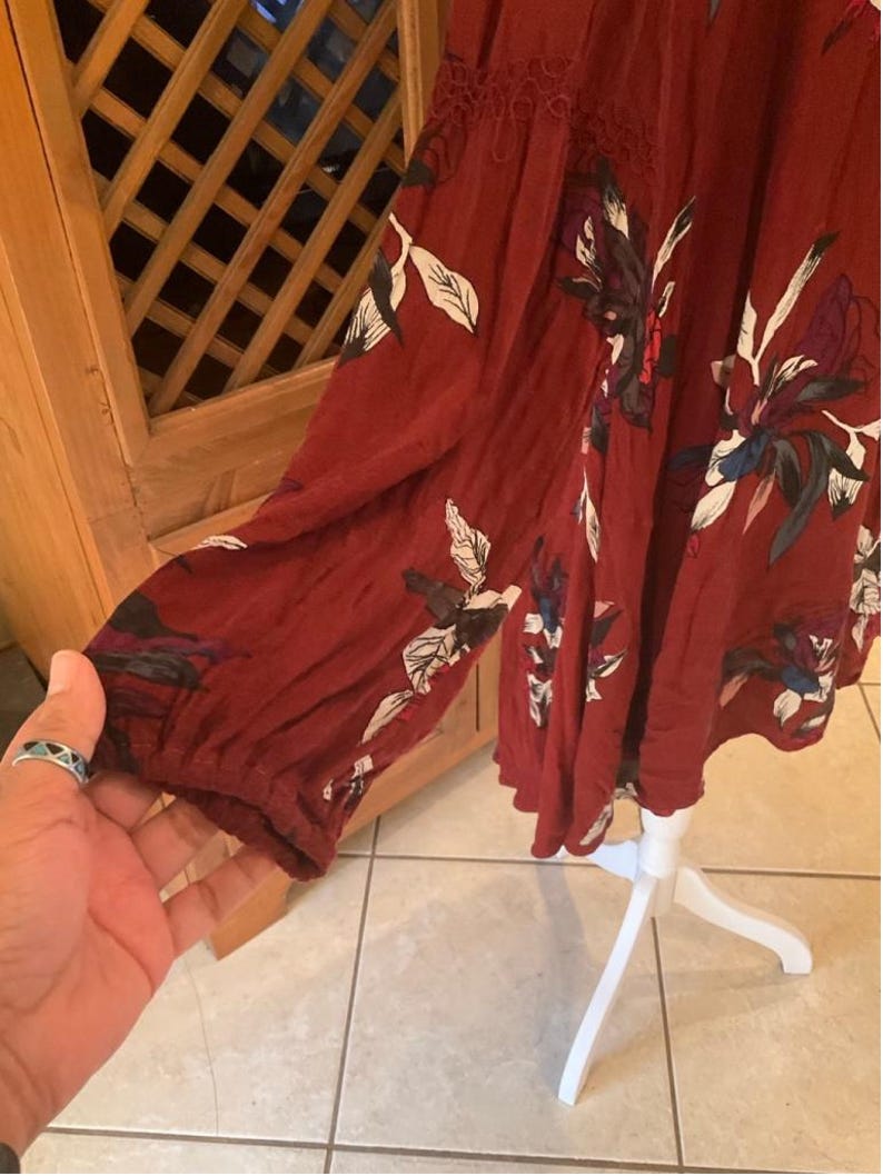 May include: A burgundy-colored dress with long sleeves and a floral pattern in white, black, and blue. The dress has a lace detail at the neckline and is displayed on a white dress form.
