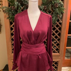 Puede incluir: Una blusa burdeos con mangas largas y cintura cruzada. La parte superior tiene un escote en V profundo y un dobladillo estilo peplum. La blusa se exhibe en un maniqu&iacute; blanco, con una corona festiva en el fondo.