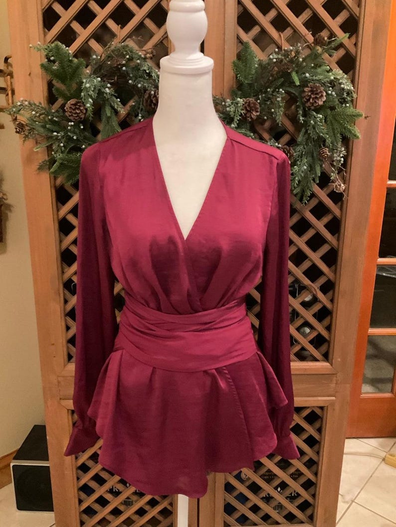 Puede incluir: Una blusa burdeos con mangas largas y cintura cruzada. La parte superior tiene un escote en V profundo y un dobladillo estilo peplum. La blusa se exhibe en un maniqu&iacute; blanco, con una corona festiva en el fondo.