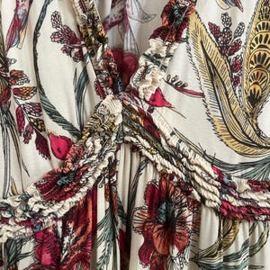 May include: Cream-coloured top with a V-neck and a floral pattern in shades of red, yellow, and green. The fabric has a ruffled trim along the neckline and a gathered detail at the centre. The design features large flowers and leaf motifs.