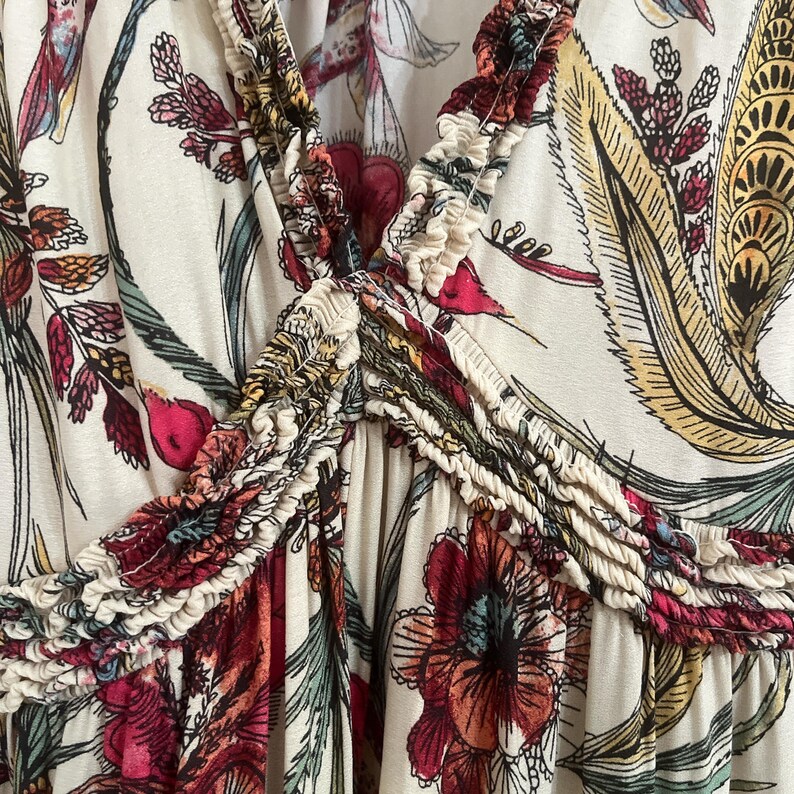 May include: Cream-coloured top with a V-neck and a floral pattern in shades of red, yellow, and green. The fabric has a ruffled trim along the neckline and a gathered detail at the centre. The design features large flowers and leaf motifs.