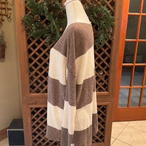 May include: A long-sleeved, oversized top with wide horizontal stripes in brown and cream. The top is displayed on a mannequin, showcasing its relaxed fit and casual style. The fabric appears soft and lightweight, suitable for layering.