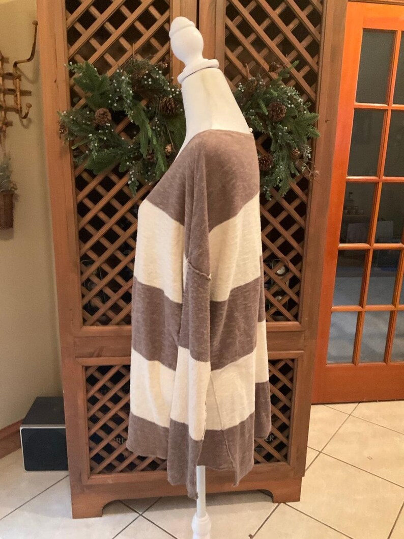 May include: A long-sleeved, oversized top with wide horizontal stripes in brown and cream. The top is displayed on a mannequin, showcasing its relaxed fit and casual style. The fabric appears soft and lightweight, suitable for layering.