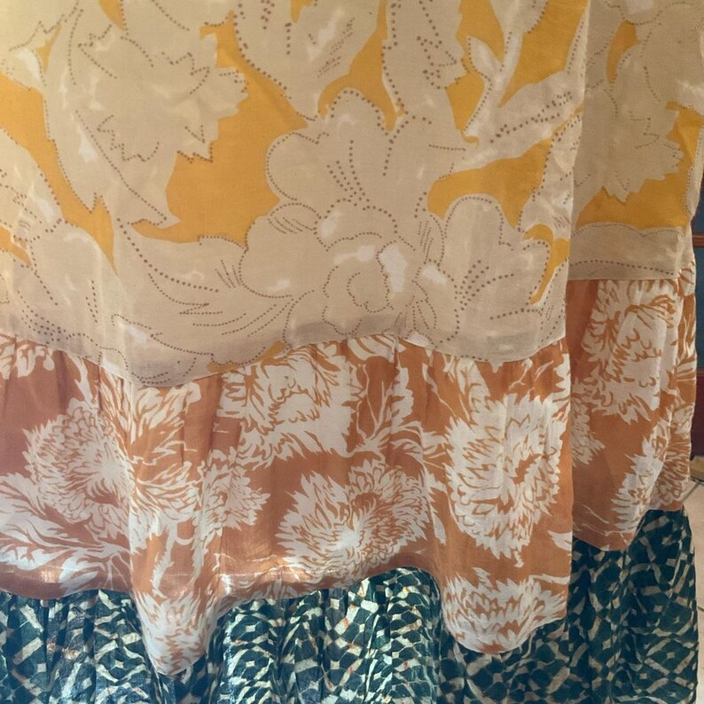 May include: Close-up of a flowing fabric with floral patterns. The top layer features large cream-colored flowers on a yellow background. Below, a band of orange and white floral print leads to a patterned blue and black bottom.