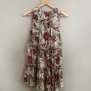 May include: Cream sleeveless dress with a floral print featuring red flowers, green leaves, and birds. The dress has a tiered skirt and is suspended on a wooden hanger. The fabric is lightweight.