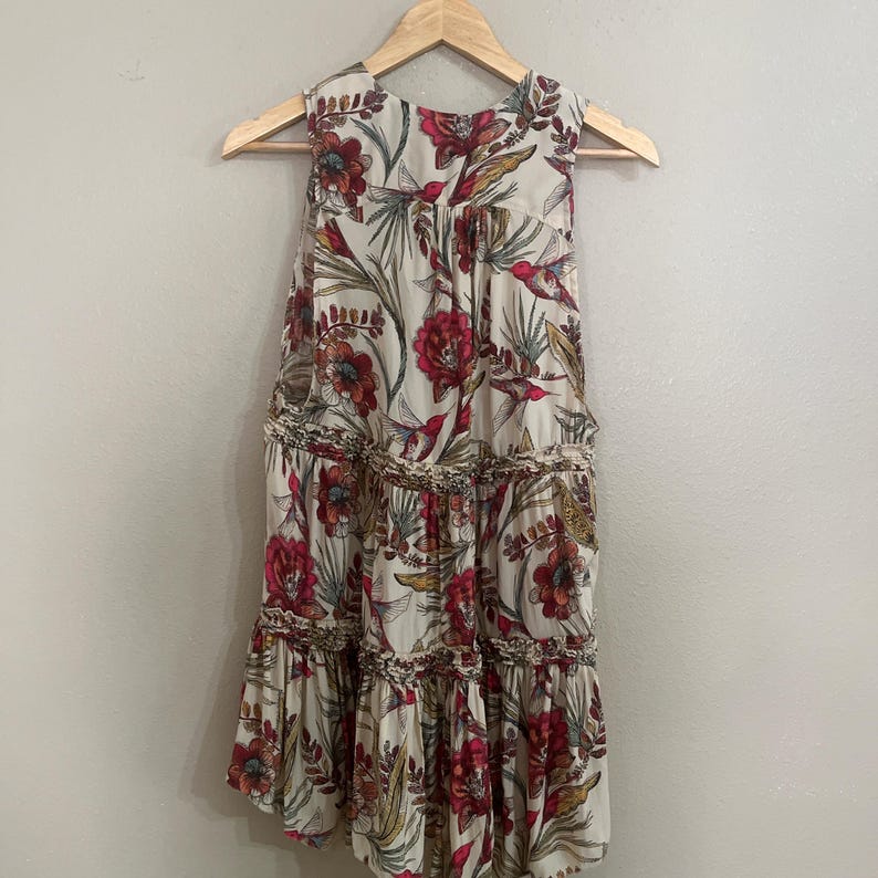May include: Cream sleeveless dress with a floral print featuring red flowers, green leaves, and birds. The dress has a tiered skirt and is suspended on a wooden hanger. The fabric is lightweight.
