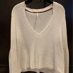 May include: Off-white knit sweater with a deep V-neck and flared sleeves. The sweater has a ribbed texture and a slightly cropped hemline. The neckline is trimmed with a contrasting burgundy fabric. The sweater is a casual, everyday garment.