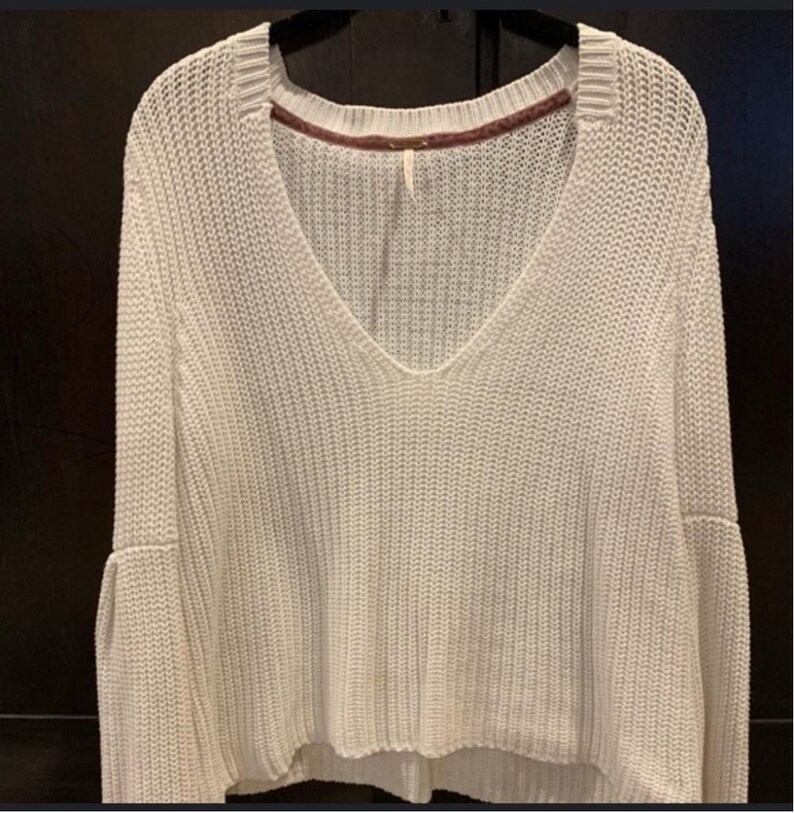 May include: Off-white knit sweater with a deep V-neck and flared sleeves. The sweater has a ribbed texture and a slightly cropped hemline. The neckline is trimmed with a contrasting burgundy fabric. The sweater is a casual, everyday garment.