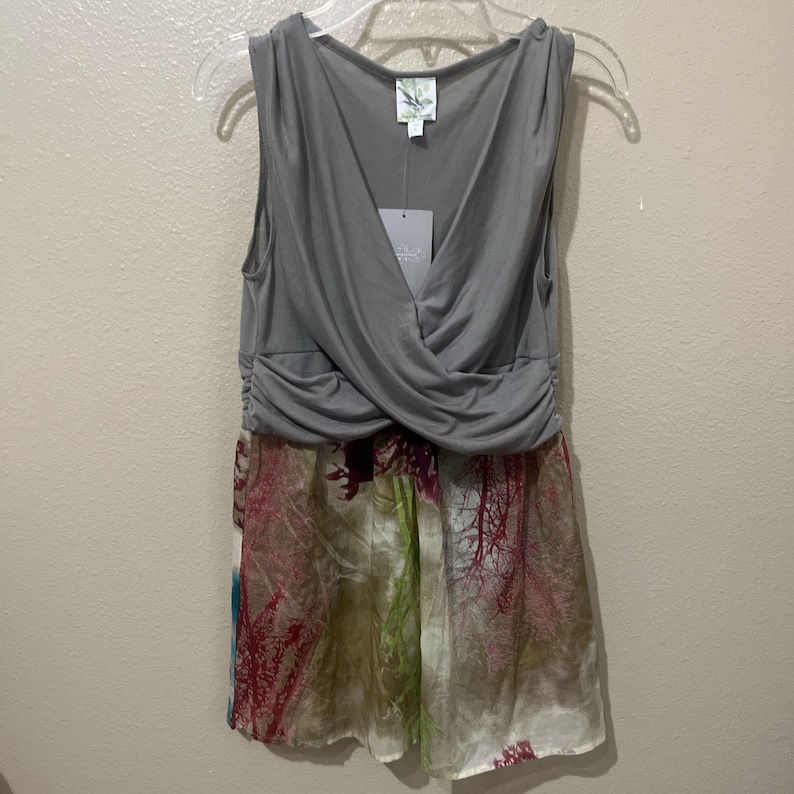 Pode incluir: Um top cinza sem mangas com um detalhe cruzado no busto, sobreposto a uma saia transparente com estampa floral. A saia tem uma base branca com estampas florais rosa, verde e marrom.
