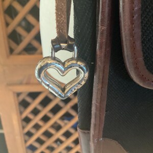 May include: Close-up of a black handbag with brown leather trim and a silver heart-shaped charm. The bag has a textured fabric body and a brown leather strap. The heart charm is attached to the strap.
