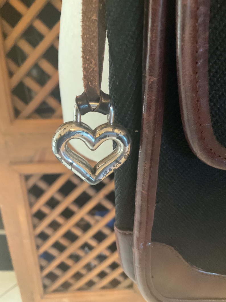 May include: Close-up of a black handbag with brown leather trim and a silver heart-shaped charm. The bag has a textured fabric body and a brown leather strap. The heart charm is attached to the strap.
