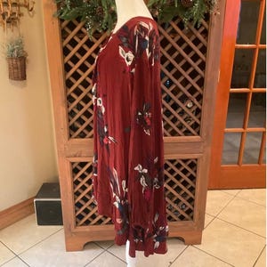 May include: A long-sleeved, burgundy dress with a floral pattern in white, red, and blue. The dress is displayed on a white mannequin. The dress has a V-neck and a ruffled hemline. The dress is made of a soft, flowing fabric.