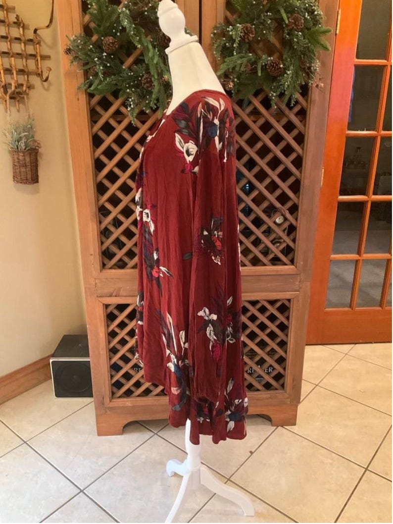 May include: A long-sleeved, burgundy dress with a floral pattern in white, red, and blue. The dress is displayed on a white mannequin. The dress has a V-neck and a ruffled hemline. The dress is made of a soft, flowing fabric.