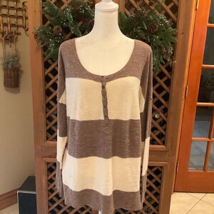 May include: A long-sleeved, scoop-neck shirt with horizontal stripes of brown and cream. The shirt has a buttoned placket and is displayed on a white mannequin. The garment is made of a soft, textured fabric.