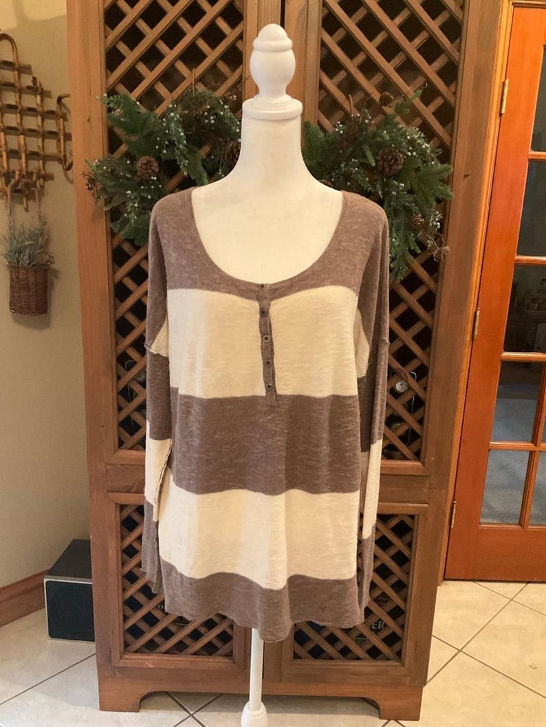 May include: A long-sleeved, scoop-neck shirt with horizontal stripes of brown and cream. The shirt has a buttoned placket and is displayed on a white mannequin. The garment is made of a soft, textured fabric.