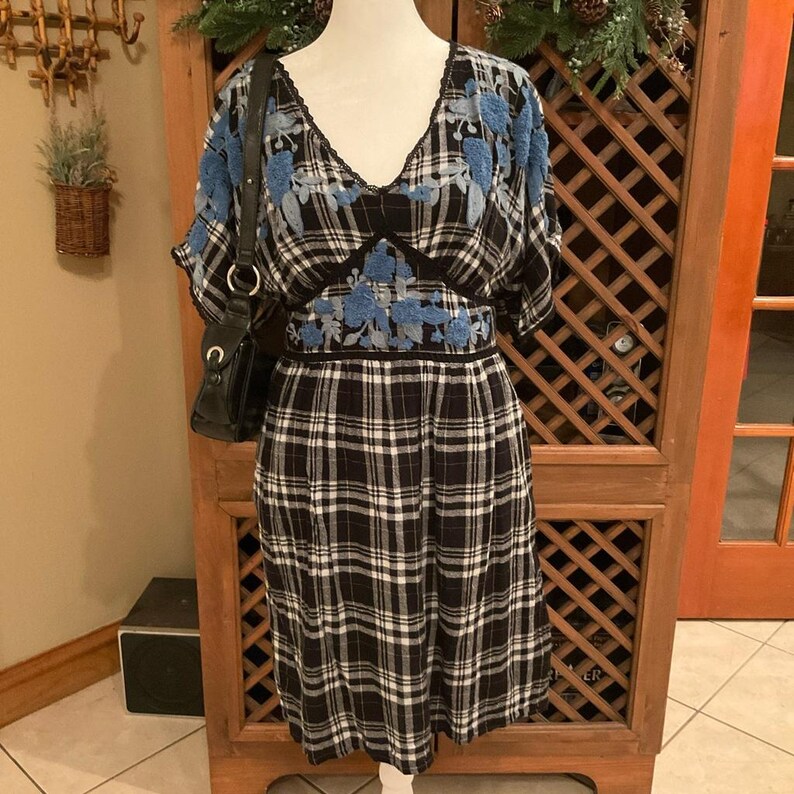 May include: A black and white plaid dress with blue floral embroidery on the shoulders and waist. The dress has a V-neck and short sleeves. A black purse hangs on the left side of the dress.