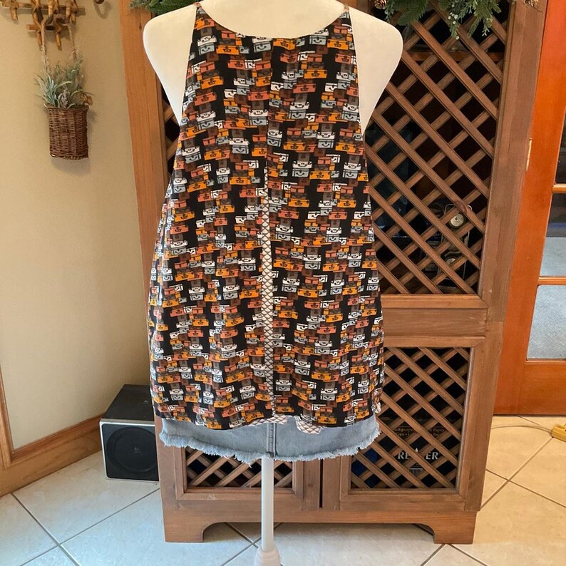Puede incluir: Una blusa sin mangas con fondo negro y un patr&oacute;n repetido de formas marrones, naranjas y blancas. La blusa tiene cuello halter y se combina con una falda vaquera.