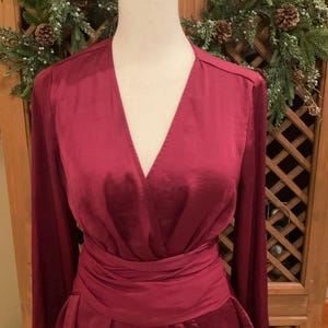 Puede incluir: Un vestido cruzado burdeos con mangas largas y cintur&oacute;n a juego. El vestido presenta un escote en V y una falda acampanada. El vestido se exhibe en un maniqu&iacute;. El fondo incluye una puerta de madera y una corona decorativa.