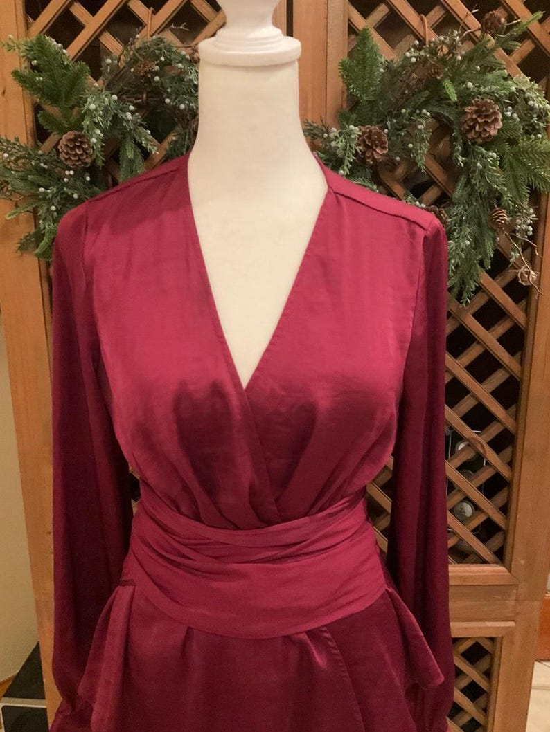 Puede incluir: Un vestido cruzado burdeos con mangas largas y cintur&oacute;n a juego. El vestido presenta un escote en V y una falda acampanada. El vestido se exhibe en un maniqu&iacute;. El fondo incluye una puerta de madera y una corona decorativa.