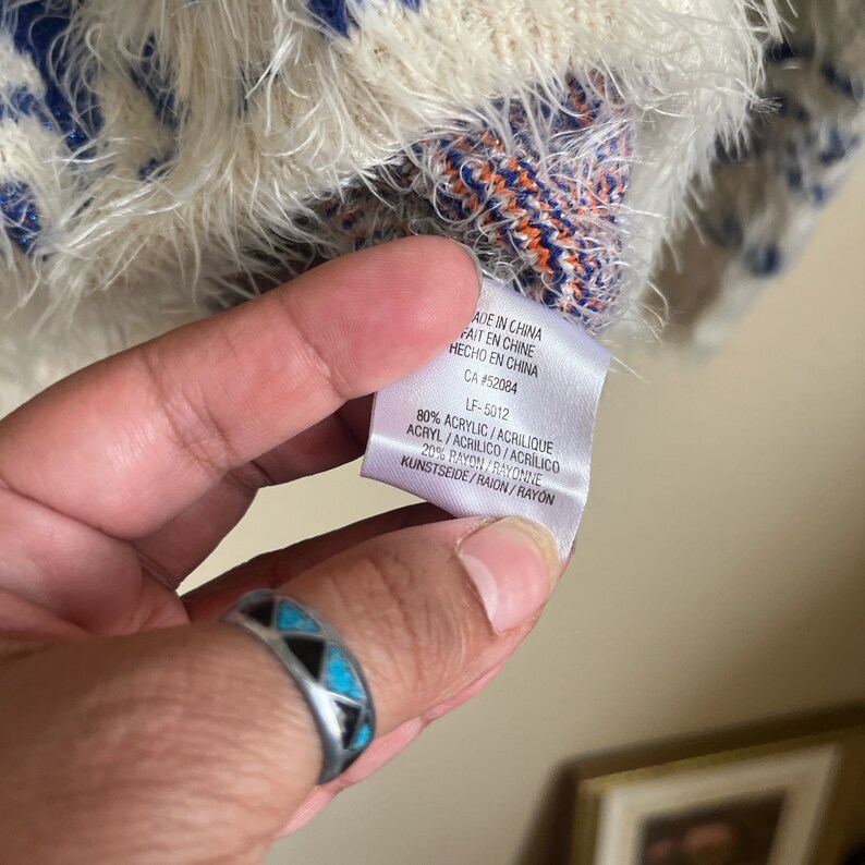 May include: A white, blue, and orange fuzzy sweater with a tag that reads "Made in China, Fait en Chine, Hecho en China, CA #52084, LF-5012, 80% Acrylic/Acrylique Acryl/Acrilico/Acrilico, 20% Rayon/Rayonne Kunstseide/Raion/Rayon".