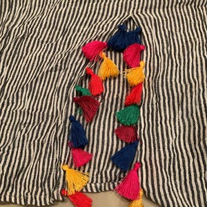 May include: Black and white striped fabric adorned with vibrant tassels. The tassels are in various colours, including red, yellow, green, and blue, and are arranged in a vertical line.