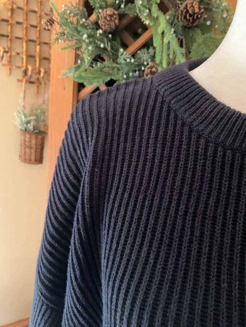 Pode incluir: Uma caneca de cer&acirc;mica branca com uma al&ccedil;a preta e o texto "But First, Coffee" em negrito preto. A caneca est&aacute; sobre um fundo desfocado, sugerindo um ambiente acolhedor.