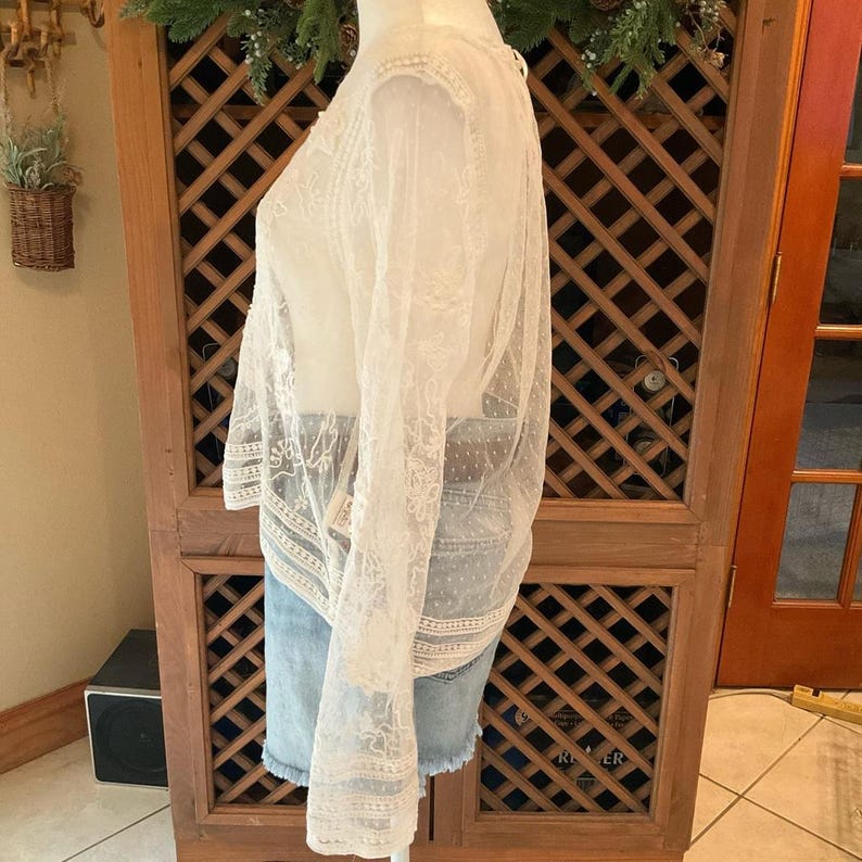 Pu&ograve; includere: Un top in pizzo bianco trasparente con maniche lunghe e intricati motivi floreali. Il top &egrave; abbinato a pantaloncini di jeans. L'indumento &egrave; esposto su un manichino, mettendo in risalto i delicati dettagli e la consistenza del pizzo.