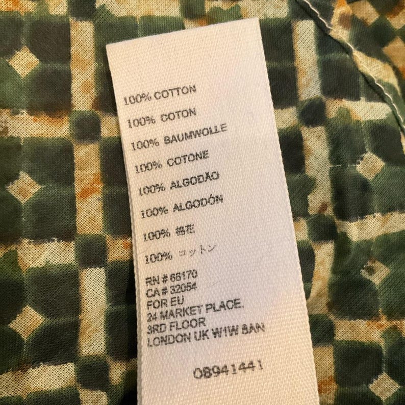 May include: A white fabric tag with text in multiple languages, including "100% COTTON" and "100% COTON." The tag also includes the address "24 MARKET PLACE, 3RD FLOOR, LONDON UK W1W 8AN" and the number "08941441."