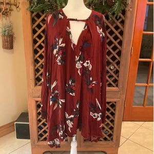 May include: A maroon long-sleeve dress with a floral pattern in white, blue, and green. The dress has a keyhole neckline and is displayed on a white mannequin. The dress is a loose, flowing style.