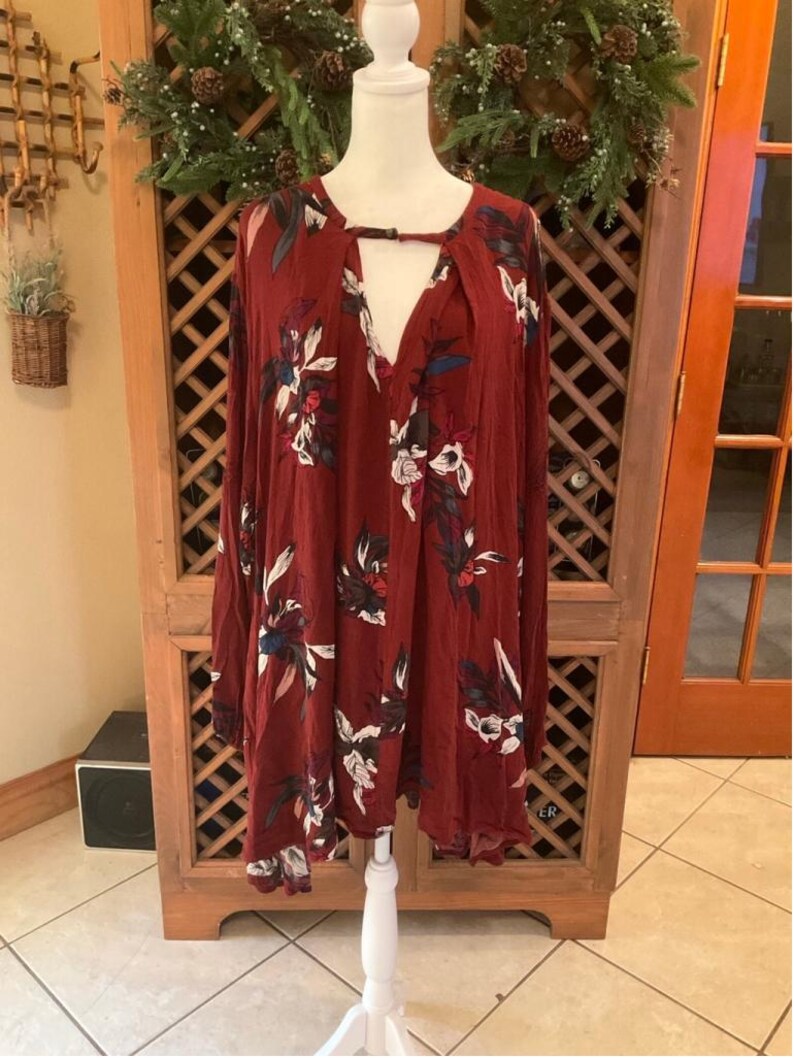 May include: A maroon long-sleeve dress with a floral pattern in white, blue, and green. The dress has a keyhole neckline and is displayed on a white mannequin. The dress is a loose, flowing style.