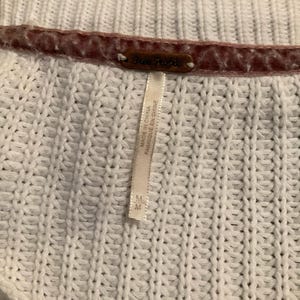 May include: Close-up of a white knit sweater with a textured pattern. The sweater has a brown tag that reads "Free People". A white tag with text is attached to the sweater. The tag reads "MADE IN CHINA FABRIQUE EN CHINE".