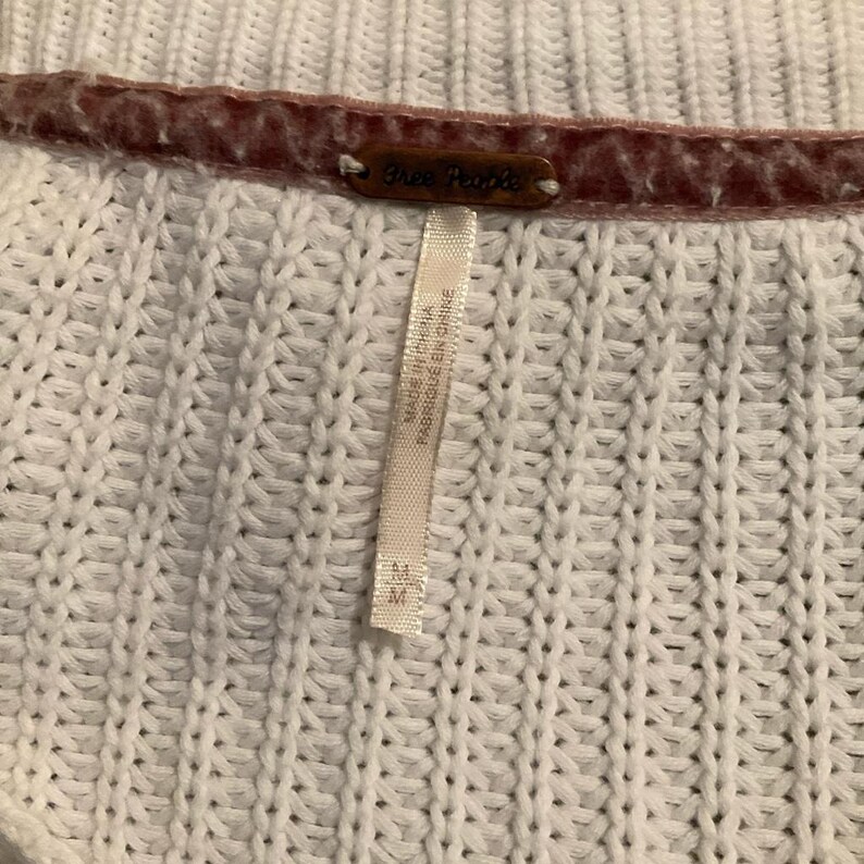 May include: Close-up of a white knit sweater with a textured pattern. The sweater has a brown tag that reads "Free People". A white tag with text is attached to the sweater. The tag reads "MADE IN CHINA FABRIQUE EN CHINE".