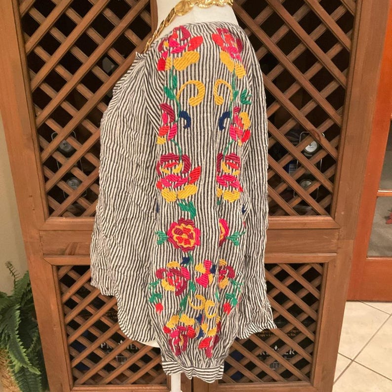 May include: A striped blouse with colourful floral embroidery. The long-sleeved top features vertical black and white stripes and vibrant embroidery in shades of red, yellow, and green. The blouse has a relaxed fit and a round neckline.