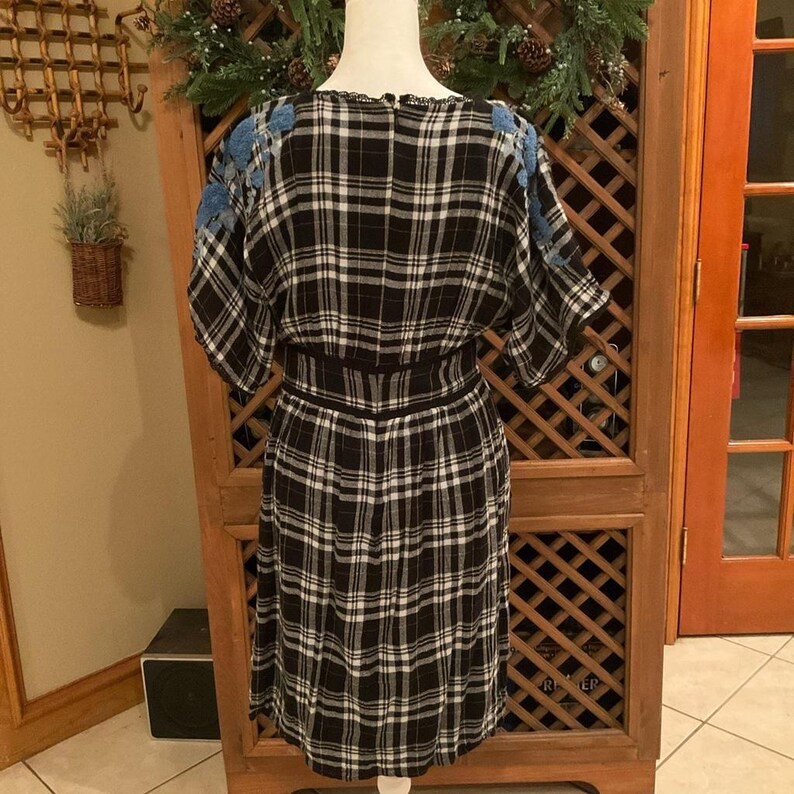 May include: A black and white plaid dress with blue floral embroidery on the shoulders. The dress has a fitted waist and a knee-length skirt. The dress is displayed on a mannequin.
